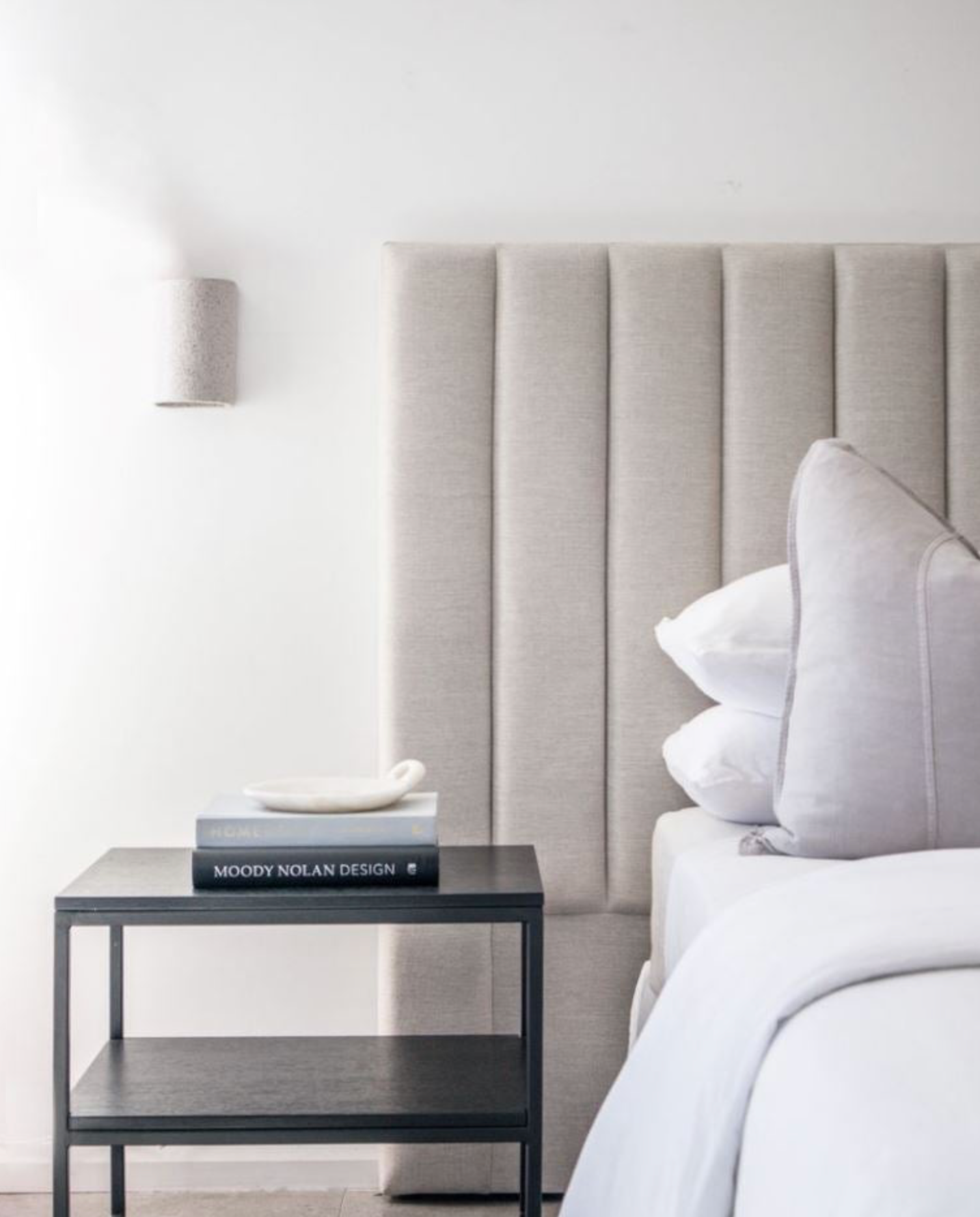 Neatly made bed with a gray upholstered headboard and a nightstand with a book and wall light in a bedroom.