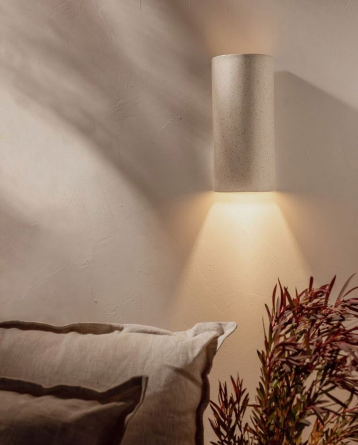 Cylindrical wall light fixture casting a warm glow on a textured wall with a pillow and plant in the foreground.