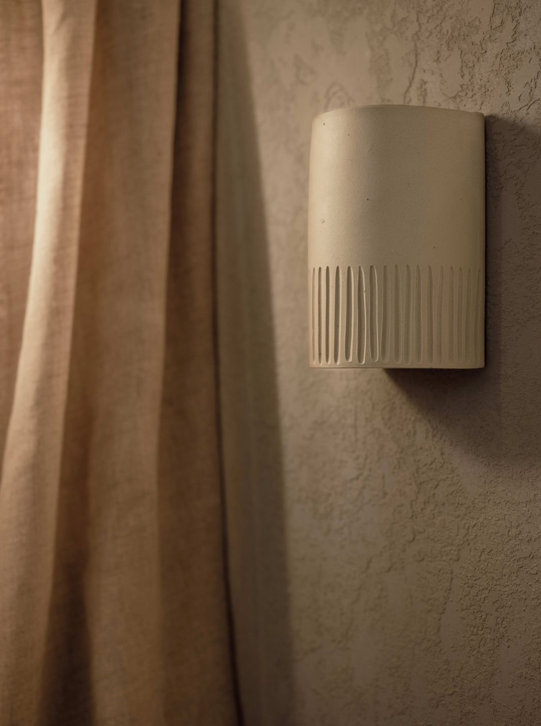 Beige wall-mounted light fixture on a textured wall with a curtain in the background